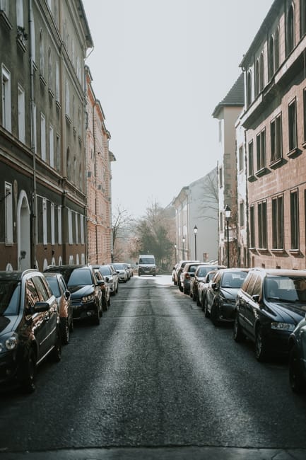 A row of parked cars along a street in Pinner during daylight, with multi-storey residential buildings in the background under a partly cloudy sky. The vehicles include various models and colours, pos