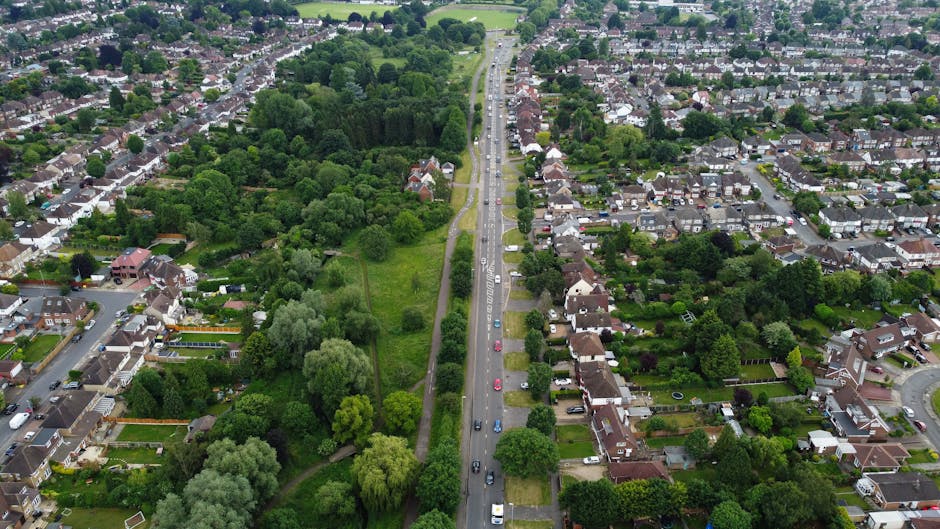 An aerial view of a residential street with a narrow road running through the centre, lined by parked cars and bordered by green gardens and trees on both sides. The street appears to be part of a suburban neighbourhood with houses and backyards visible in the background. In the foreground, a removals team from Pinner Man and Van is engaged in a house relocation effort, with a moving van parked on the pavement and furniture wrapped in protective fabrics and cardboard boxes being loaded or unloaded. Trolleys and straps are being used to facilitate the transportation of household goods, and the scene captures the logistics of furniture transport and packing during the home moving process. The surrounding environment includes lush greenery, well-maintained gardens, and a mix of brick and tiled houses, illustrating a typical neighbourhood setting for professional removals services focused on house moves and relocation logistics.