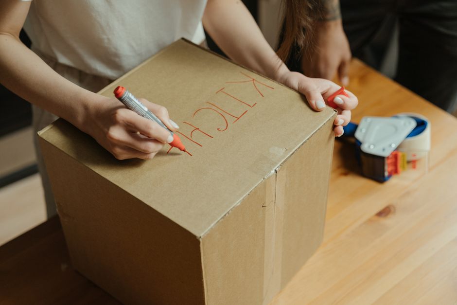 Several cardboard moving boxes of various sizes are stacked on a light wooden floor inside a home. The boxes are sealed with packing tape, and some have black plastic handles on their sides. In the ba