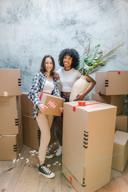Two women inside a room during a house removal process, surrounded by numerous cardboard boxes sealed with red tape and stacked nearby. One woman, wearing a checked shirt and beige trousers, holds a medium-sized box. The other woman, dressed in a white top and plaid trousers, carries a white woven basket with large green tropical leaves sticking out. The room has a textured grey wall and a wooden floor with some packing material scattered around. The women are smiling and appear to be coordinating as part of a furniture transport or packing and moving service provided by Pinner Man and Van, with the scene capturing a moment in their home relocation or urgent move. A van may be nearby, but it is not visible in this image.