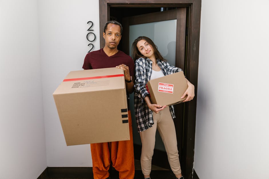 Two women inside a room during a house removal process, surrounded by numerous cardboard boxes sealed with red tape and stacked nearby. One woman, wearing a checked shirt and beige trousers, holds a medium-sized box. The other woman, dressed in a white top and plaid trousers, carries a white woven basket with large green tropical leaves sticking out. The room has a textured grey wall and a wooden floor with some packing material scattered around. The women are smiling and appear to be coordinating as part of a furniture transport or packing and moving service provided by Pinner Man and Van, with the scene capturing a moment in their home relocation or urgent move. A van may be nearby, but it is not visible in this image.