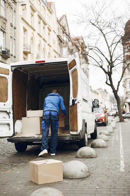 A male removal service worker with a beard and short hair, dressed in a navy and white uniform, is sitting at the side of a white moving van with the sliding door open. He is holding a clipboard and a